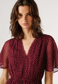 Vestido burdeos con mangas cortas transparentes, estampado de lunares, escote en V profundo, escote deshilachado y cintura fruncida para una silueta fluida.