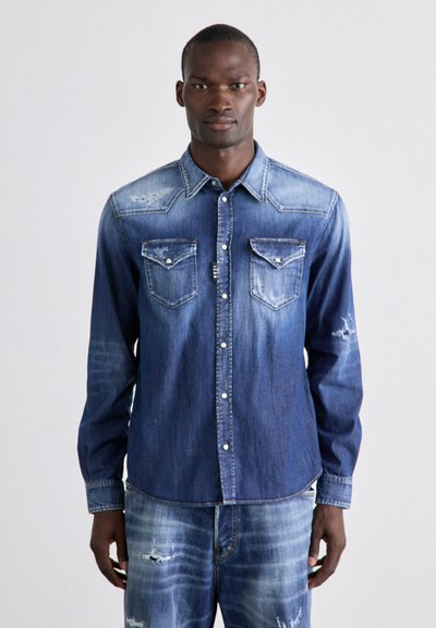 Chemise en denim bleu délavé avec deux poches poitrine, fermeture à boutons à l'avant et détails usés. Manches longues avec poignets à bouton.