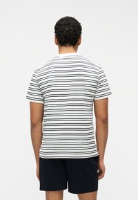 Άνδρας με κοντά σγουρά μαλλιά, φοράει λευκό T-shirt με μαύρες οριζόντιες ρίγες και μαύρο σορτς, στέκεται γυρισμένος με την πλάτη σε απλό φόντο.