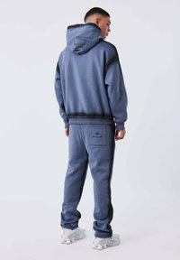 Hombre con sudadera con capucha azul y pantalones deportivos a juego con logo en el bolsillo, vistiendo zapatillas blancas, de pie contra un fondo claro y liso.