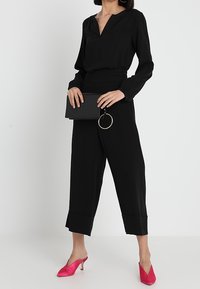 Sort langærmet bluse med V-udskæring, højtydende sorte vide bukser og lyserøde spidse sko, mens der holdes en sort clutch med en cirkulær accent.