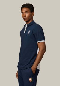 Man draagt een marineblauw-witte poloshirt en bijpassende broek met Hackett London Rowing Club-logo's, staand met handen in zakken.