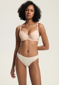 Kobieta o kręconych włosach, ubrana w jasnobeżowy koronkowy stanik i kremowe majtki, stojąca z jedną ręką za plecami na jednolitym tle.