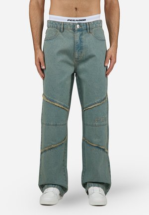 Helle blaue Denim-Hose mit lockerer Passform, die fransige diagonale Akzente und Logo-Stickerei aufweist. Mit einem weißen Bund gestylt.