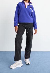 Pullover in pile viola con accenti rossi, colletto bianco e tasca con zip, abbinato a jeans neri larghi e sneakers bianche.