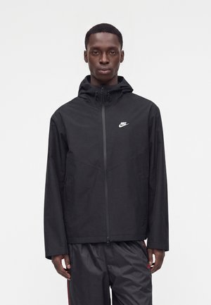 Мъж, облечен в черно яке с качулка на Nike с преден цип и черен панталон, застанал пред бял фон.
