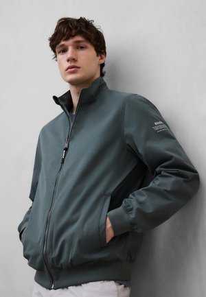 Joven con chaqueta de cremallera verde oscuro y manos en los bolsillos, de pie contra una pared gris claro, mirando a la cámara.