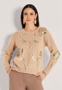 Frau, die einen beigen Pullover mit goldener floraler Paillettenbestickung und braunen Hosen trägt, steht mit den Händen in den Taschen vor einem neutralen Hintergrund.