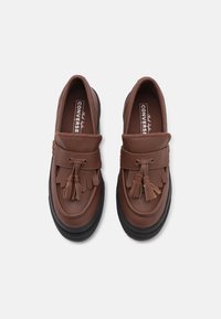 Brune skinnloafers med rund tå, dekorativ frynse og dusk, samt en teksturert svart gummisåle. Converse-merke på innersålen.