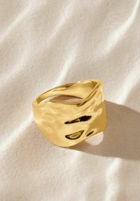 Goldring mit einem welligen, skulpturalen Design und polierter Oberfläche. Glatte Textur, sich verjüngende Form, die Licht gegen einen sanften Hintergrund reflektiert.