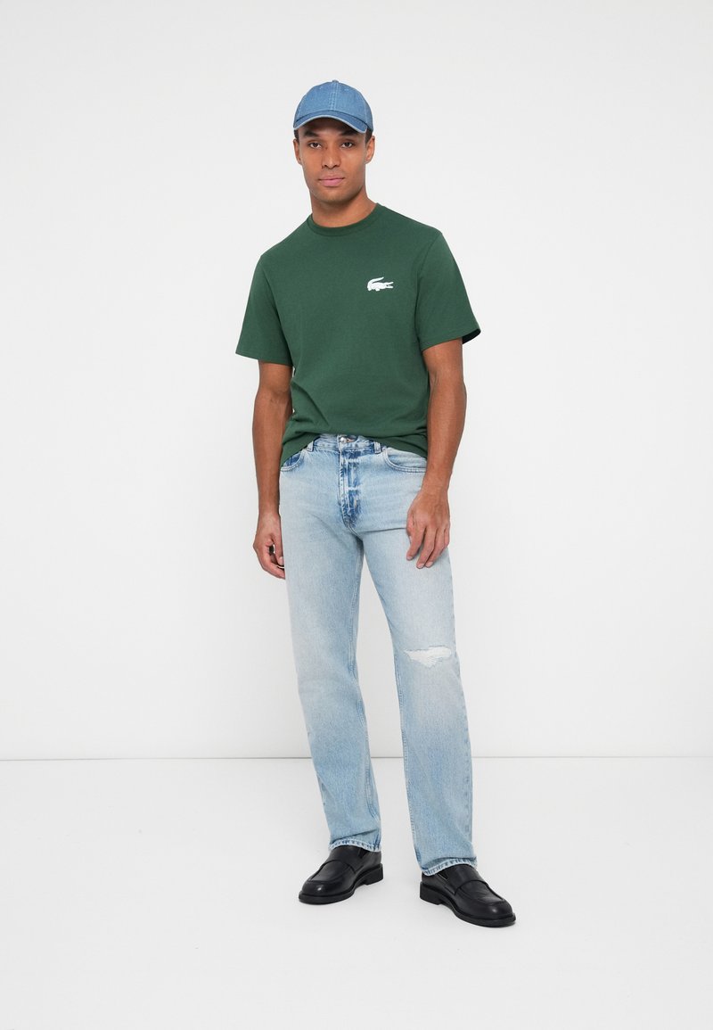 Grünes Baumwoll-T-Shirt mit Lacoste-Logo, hellblaue, ausgebleichte Jeans und schwarze Slipper, kombiniert mit einer hellblauen Baseballkappe.