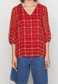 Blusa roja con estampado, escote en V profundo, mangas abullonadas y diseño a cuadros. Hecha de tela ligera, combinada con pantalones cortos de mezclilla azules.
