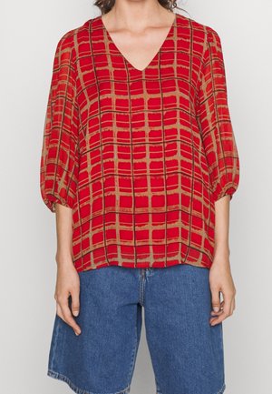 Blusa rossa con motivo a fantasia, scollatura a V profonda, maniche a palloncino e design a griglia. Realizzata in tessuto leggero, abbinata a pantaloni corti in denim blu.