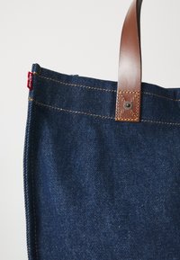 Denim tas met een donkerblauwe kleur, bruine leren handgreep en contrasterende gele stiknaad. Heeft een rode label aan de zijkant.
