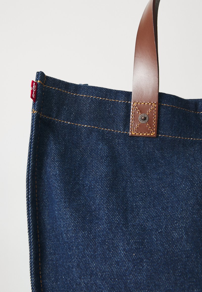 Denim tas met een donkerblauwe kleur, bruine leren handgreep en contrasterende gele stiknaad. Heeft een rode label aan de zijkant.