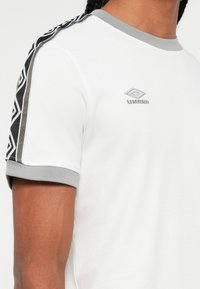 Hvit kortermet skjorte med grå krage og mansjetter, svart og grå geometrisk mønster på skuldrene, og grå Umbro-logo på brystet.