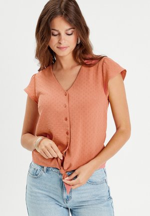 Blouse à manches courtes et col en v couleur orange rouille avec un motif texturé. Présente des boutons au centre et un détail noué à l'ourlet.