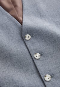 Tissu gris clair à texture tissée, avec trois boutons argentés et une doublure intérieure marron visible à côté du col.