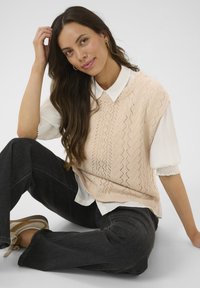 Gilet en tricot pêche avec un motif en losange, superposé à une chemise blanche à col. Un jean noir à jambes larges et des chaussures beiges complètent la tenue.