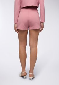 Pantaloni shorts rosa su misura realizzati in un tessuto liscio con una vita aderente. Indossati con scarpe con tacco chiare, che mostrano un design semplice.