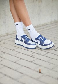 Vita och marinblå Nike-sneakers med en texturerad läderöverdel, platt vit sula och blå snörningar, bärs med vita ankelsockor.