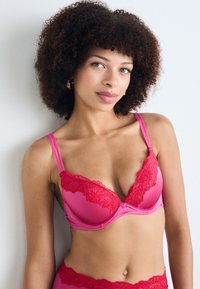 Roze beha met kanten rand en een diep uitgesneden halslijn, voorzien van verstelbare bandjes. Glad materiaal in een levendige kleur, met bloemige kantaccenten.