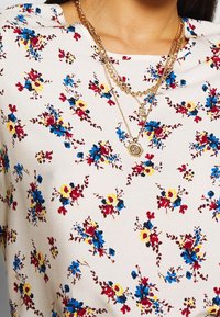 Blommig blus i lätt material med en krämfärgad bakgrund och mönster av röda, gula och blå blommor, lager på lager med guldtonade halsband.