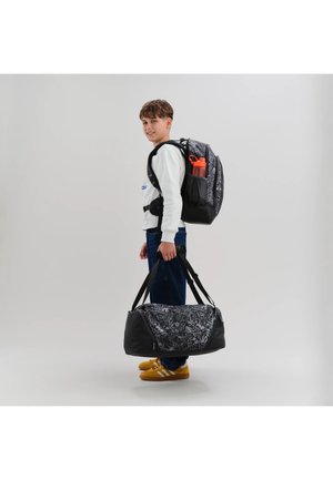 Ragazzo in abbigliamento casual che porta una borsa da viaggio nera con motivo e indossa uno zaino abbinato con una borraccia arancione nella tasca laterale.