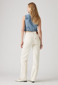 Γυναίκα με μακριά ξανθά μαλλιά που φοράει αμάνικο μπλε τζιν γιλέκο, λευκά τζιν ψηλόμεσα και λευκά παπούτσια με χαμηλό τακούνι, στεκόμενη με την πλάτη γυρισμένη.