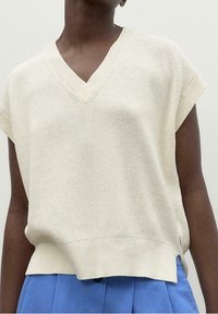 Light beige knitted sweater with a V-neck and short sleeves. Smooth texture, loose fit, and a straight hemline. Worn over blue trousers.