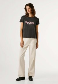 T-shirt preta com "Pepe Jeans London" em texto rosa, combinada com calças largas bege e sapatos pretos. Design simples, ajuste casual e textura suave.