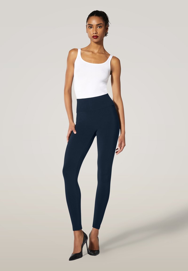 Högmidjade marinblå leggings i stretchtyg, matchade med en vit tanktop och svarta klackar, stående på en ljus bakgrund.