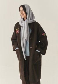 Barna gyapjúkabát két első zsebbel, hímzett alma és virág díszítéssel, világoskék sállal és alatta egy pulóverrel párosítva.