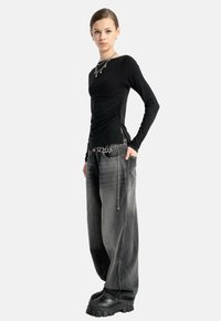 Młoda osoba w dopasowanej czarnej bluzie z długim rękawem, szerokich, wyblakłych czarnych dżinsach, masywnych czarnych butach oraz srebrnej biżuterii i pasku z motywem gwiezdnym.