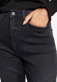 Jeans in denim nero dal design aderente con applicazioni di strass sparsi. Include una tasca frontale e chiusura con bottone metallico.