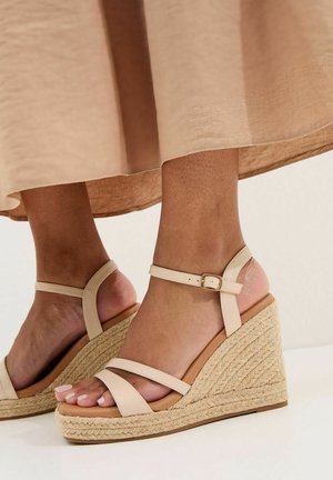 Sandalias de cuña tipo alpargata en color marrón claro con correas al tobillo y hebillas doradas, usadas con una falda beige, mostrando los pies y las piernas inferiores.