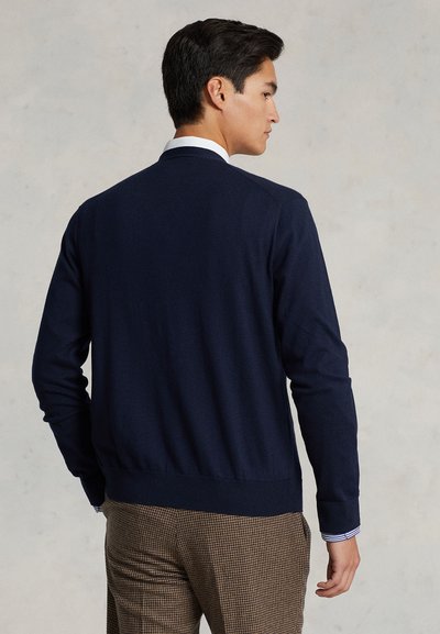 Maglione a maglia blu navy con maniche lunghe e orlo a costine. Indossato sopra una camicia bianca con colletto caratterizzata da strisce blu.