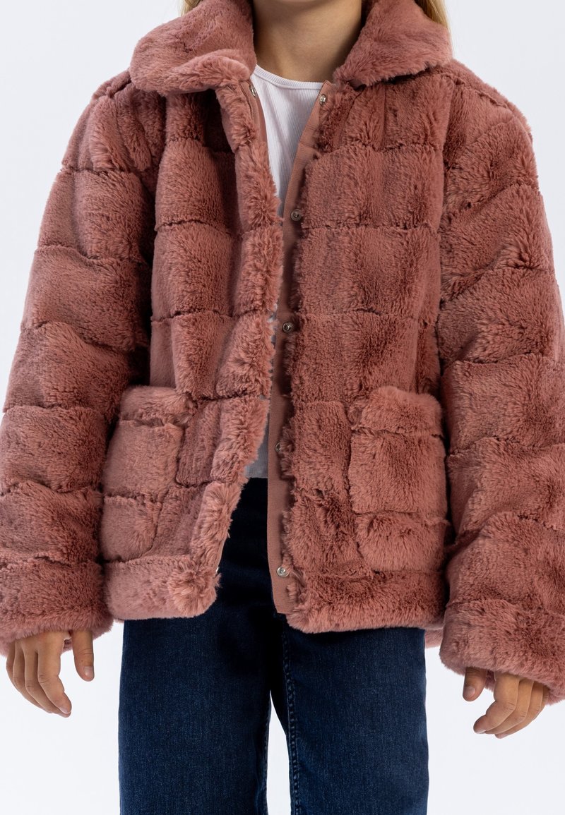 Personne portant une veste matelassée en fausse fourrure rose poussiéreuse avec un grand col et des poches, associée à un jean bleu foncé et une chemise blanche en dessous.