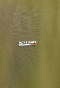 Λογότυπο JACK & JONES σε λευκό πάνω από μια μικρή οριζόντια γραμμή σε μπλε, λευκό και κόκκινο χρώμα σε επιφάνεια ελιάς ύφασμα.