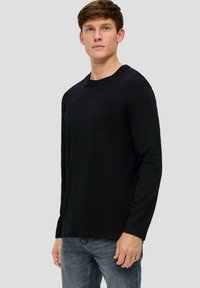 Schwarzes Langarmshirt aus weichem Stoff, mit Rundhalsausschnitt, Brusttasche und lockerer Passform, kombiniert mit hellblauen Jeans.