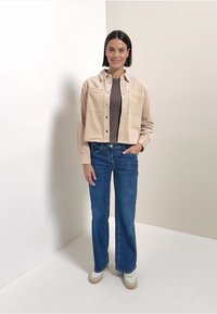 Chaqueta de pana beige con cierre de botones, llevada sobre una blusa marrón, combinada con jeans de campana de color azul claro y zapatillas blancas. Textura suave.