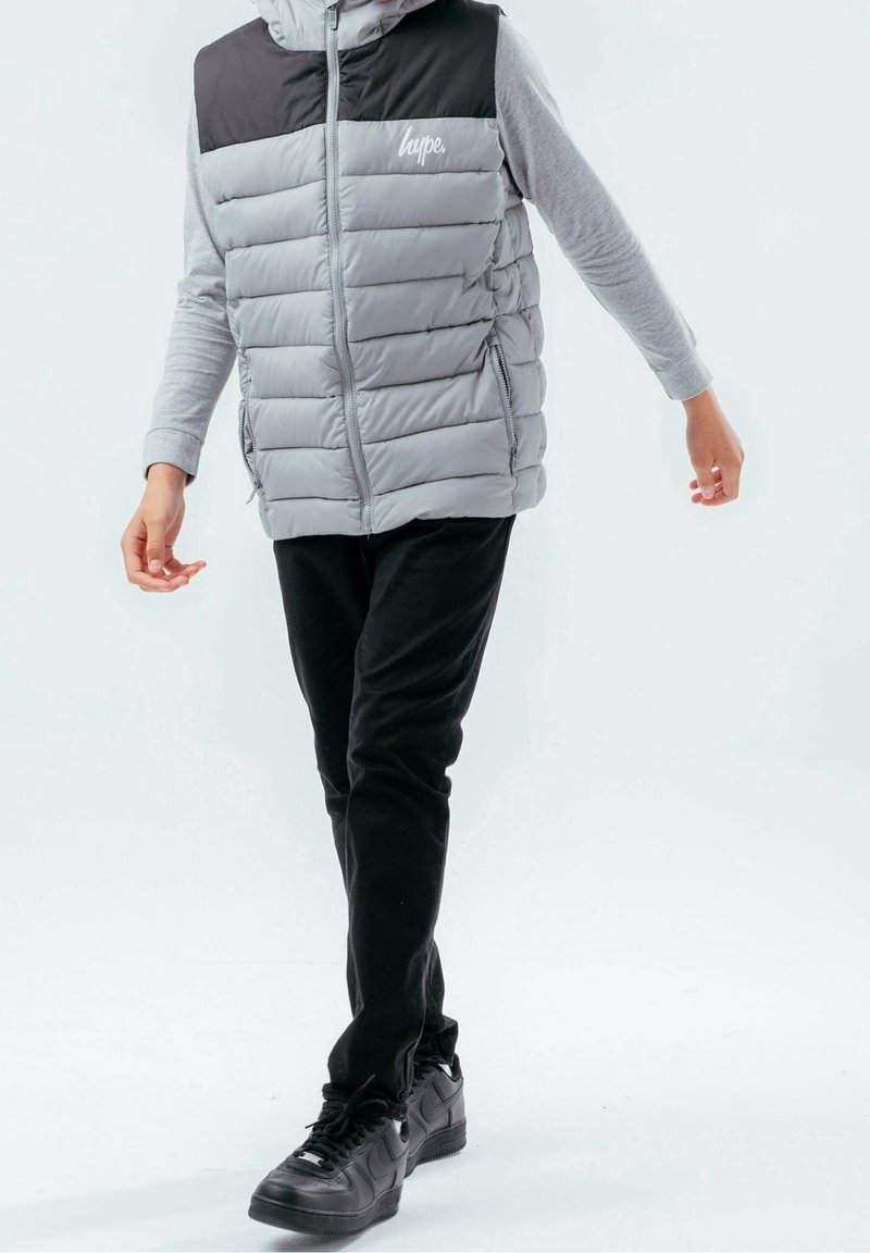 Veste grise matelassée avec épaules noires, fermeture éclair, logo sur la poitrine, assortie à un pantalon noir et des baskets noires. Texture plate, coupe décontractée.