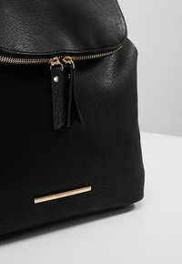 Sac à dos en cuir noir texturé avec fermeture éclair dorée et doubles tirettes en cuir, équipé d'une petite plaque nominative horizontale en or.