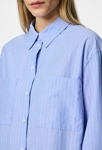 Camicia azzurro chiaro con righe verticali bianche, dotata di colletto e due tasche sul petto. Realizzata in un tessuto morbido con una vestibilità comoda.