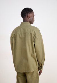 Olive green oversized shirt made of cotton with a pointed collar, long sleeves, and buttoned cuffs. The back has a yoke seam detail.