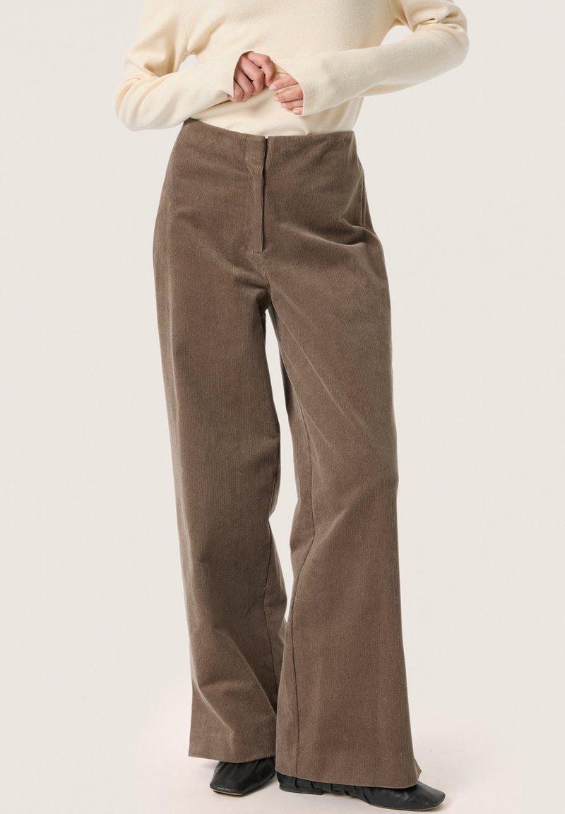 Brown corduroy wide-leg trousers with a high waist and textured fabric, paired with a light-colored long-sleeve top.