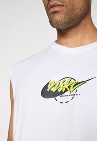Közelkép egy férfiról, aki fehér ujjatlan pólót visel, rajta egy sárga és zöld "Nike Basketball" logóval, amely egy fekete pipa design felett helyezkedik el.