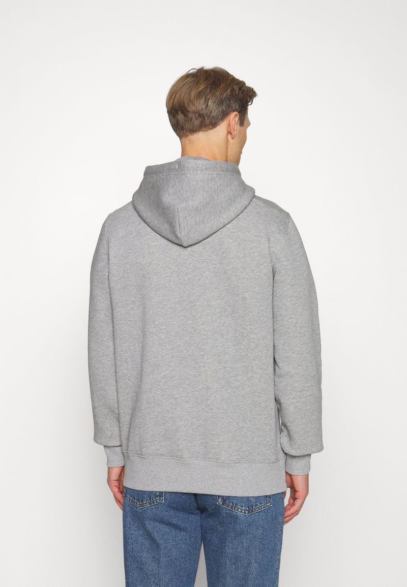 Hombre con cabello corto marrón, vistiendo una sudadera gris lisa y jeans azules, mostrado de espaldas sobre un fondo blanco.