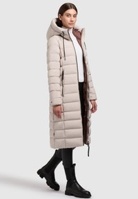Dlhý béžový zipsový kabát s kapucňou, s prešívaným dizajnom, zipsovým zapínaním a čiernymi akcentmi, skombinovaný s čiernymi členkovými topánkami.