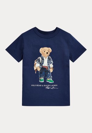 Camiseta de algodón azul marino con un gráfico de un oso en ropa casual, incluyendo un chaleco estampado, vaqueros y zapatillas verdes.